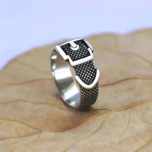 Silver-tone ring shaped like a belt with a buckle and a dark textured band, resting on a leaf
