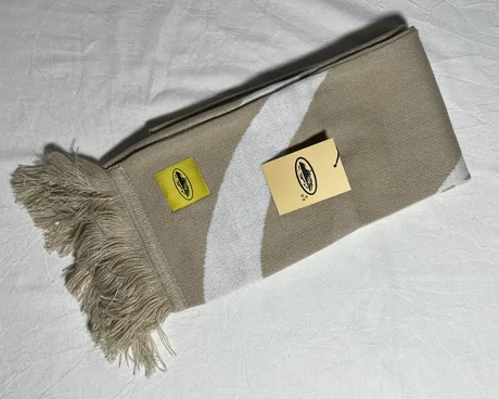 Khaki knit scarf laid flat with a fringed end and a small woven badge